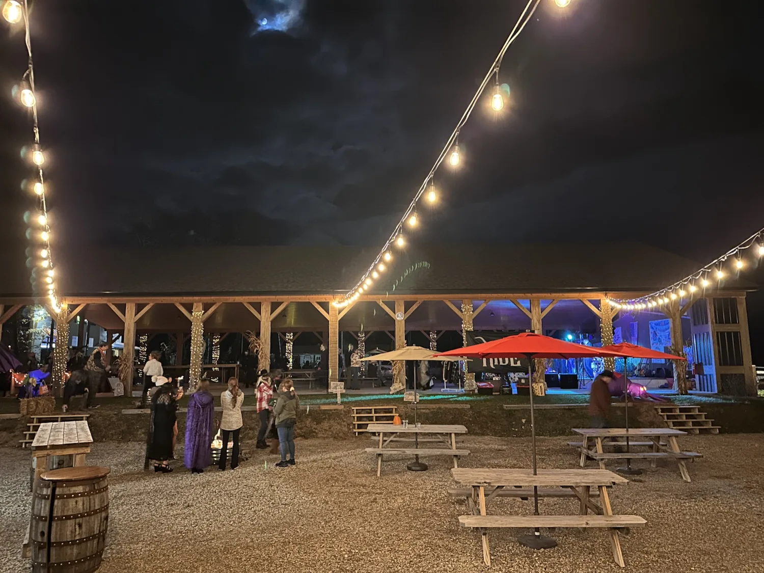 Wide night view from the backyard patio under string lights looking into the lit timber-frame pavilion, with guests around a fire pit and picnic tables