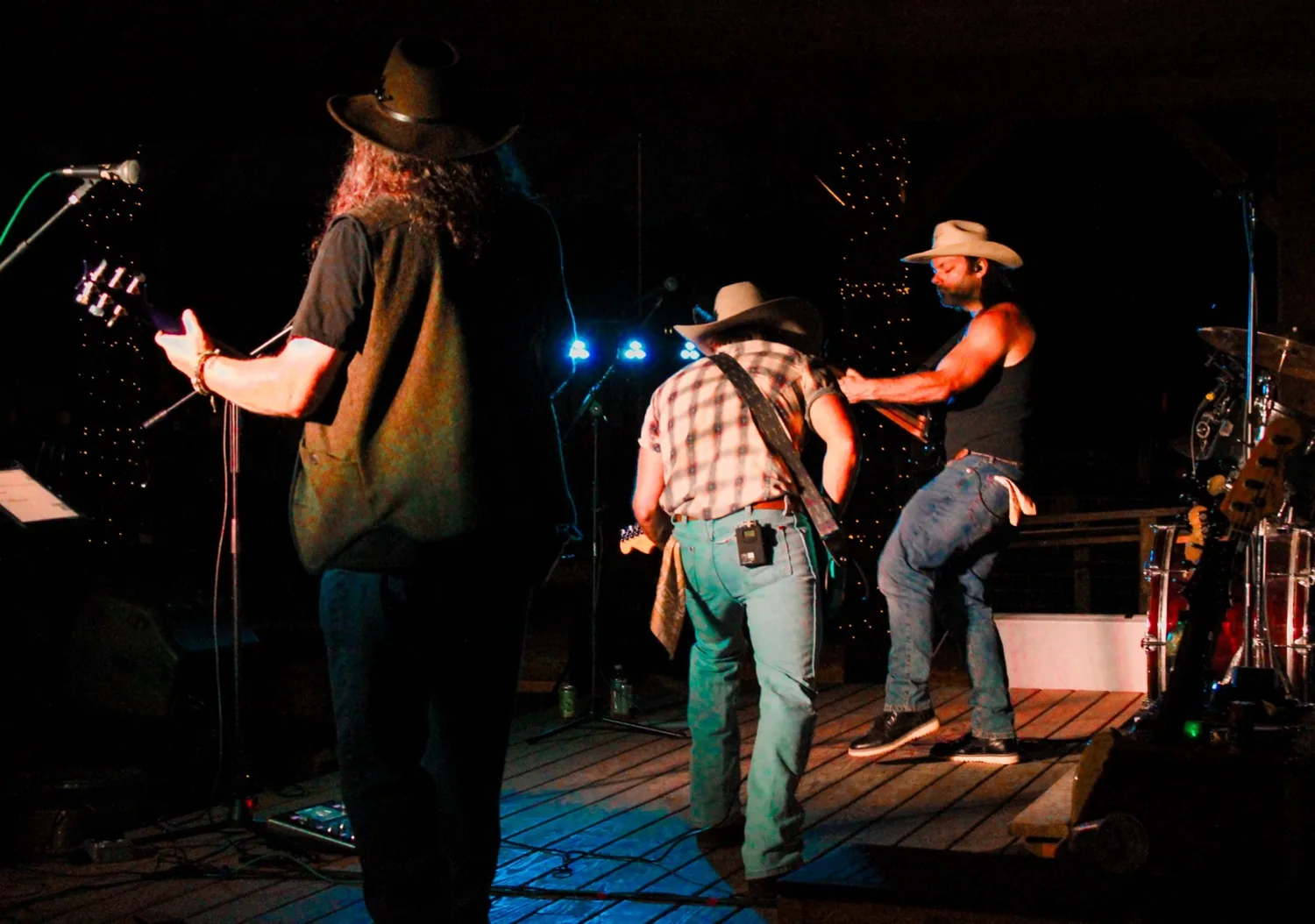Three musicians in cowboy hats playing guitars on the wooden pavilion stage under blue and white stage lights