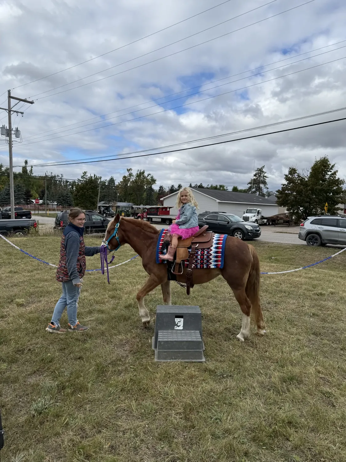 Child in a pink skirt riding a saddled pony led by a handler in the fenced field at the 2nd Annual Harvest Festival