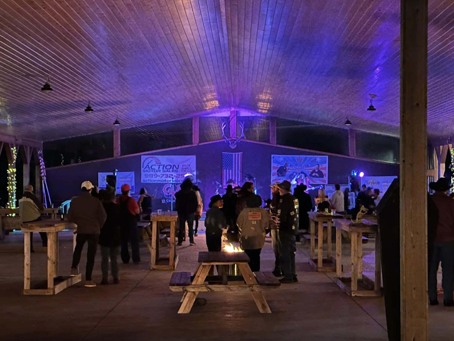 Inside the timber-frame pavilion at night during a live concert with purple stage lighting on the ceiling, high-top tables and a fire table