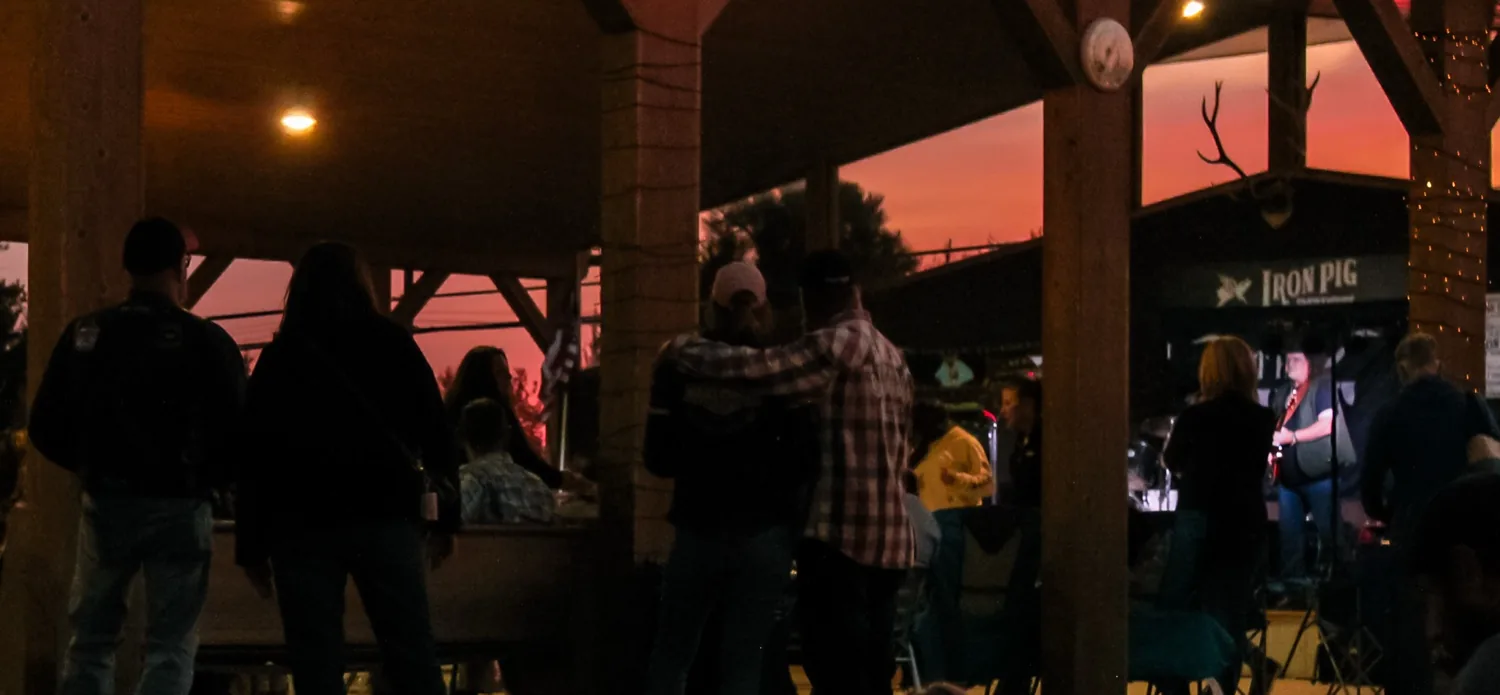 Couples and guests under the timber pavilion at sunset watching a live band on stage with a pink and orange sky behind the rafters
