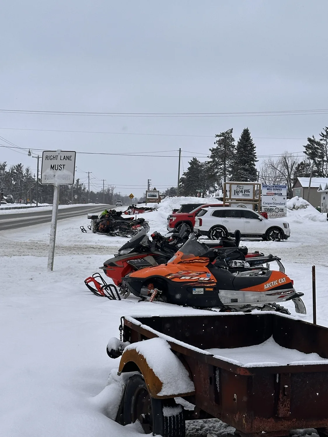 Row of Arctic Cat and Ski-Doo snowmobiles parked in the snow along Old US Hwy 27 S in front of the Iron Pig Smokehouse road sign