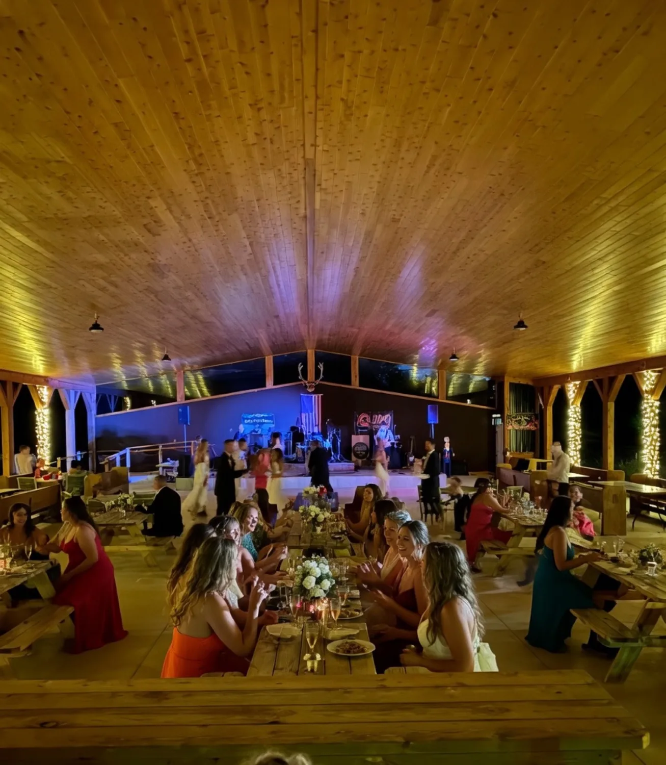 Inside the timber-frame pavilion at night during a wedding reception dinner with long wooden banquet tables, white floral runners, candlelight and guests in evening dresses