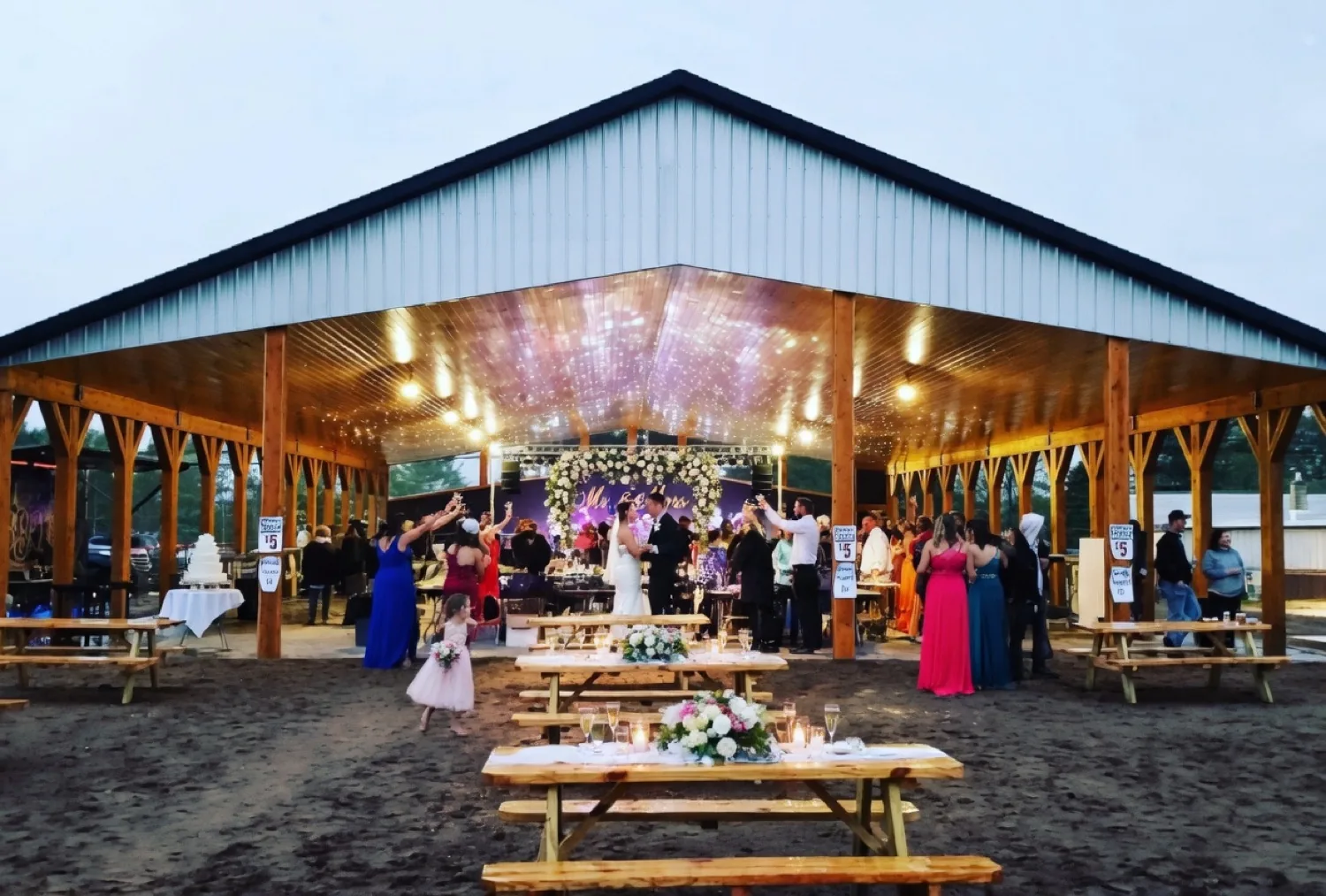 Bride and groom dancing under a white floral arch and Mr. and Mrs. sign inside the lit timber-frame pavilion while guests raise their glasses